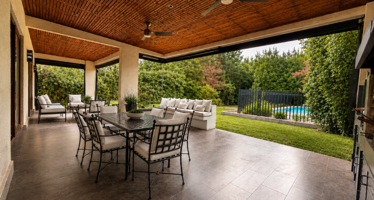 Terraza techada con comedor exterior, zona de descanso, jardín y piscina al fondo.