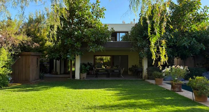 Vista posterior de la casa con amplia terraza techada, jardín y árboles consolidados.